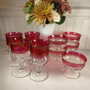 Può includere: Una collezione di bicchieri vintage rosso rubino, tra cui calici e piccole coppette da dessert. I bicchieri hanno steli trasparenti a motivi e bordi con finiture dorate. Un vaso bianco con fiori sullo sfondo.