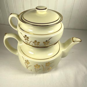 May include: A cream-colored ceramic tea for one set consisting of a teapot base and a stacking lid-topped cup. The set features a delicate gold floral pattern and gold trim along the rims, handle, and spout, creating a cohesive two-piece design.