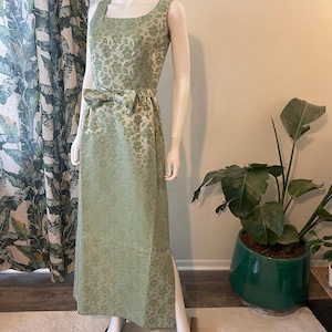 May include: A sleeveless, floor-length, light green dress with a floral pattern. The dress features a square neckline, a bow at the waist, and a slit on one side. The dress is displayed on a mannequin.