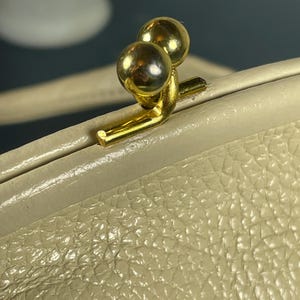 May include: Close-up of a cream-colored purse clasp with a textured leather exterior. The clasp features two gold-colored spheres and a gold-colored metal frame. The purse appears to be vintage.