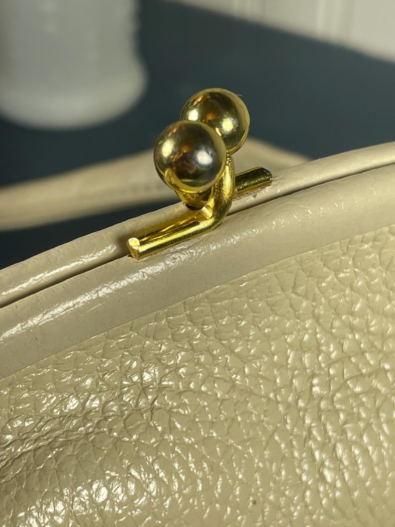 May include: Close-up of a cream-colored purse clasp with a textured leather exterior. The clasp features two gold-colored spheres and a gold-colored metal frame. The purse appears to be vintage.