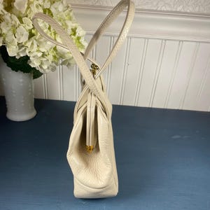 May include: Cream-colored vintage handbag with a structured design. The purse features a clasp closure and dual handles. The bag is made of a textured material and has gold-toned hardware. A classic accessory for any occasion.