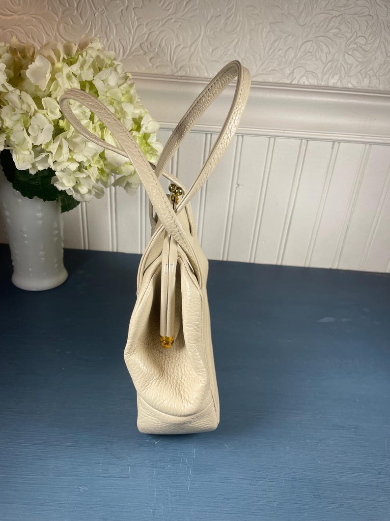May include: Cream-colored vintage handbag with a structured design. The purse features a clasp closure and dual handles. The bag is made of a textured material and has gold-toned hardware. A classic accessory for any occasion.