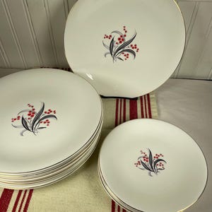 Puede incluir: Un conjunto de platos de cerámica blanca con borde dorado y un diseño floral con hojas negras y bayas rojas. Los platos están apilados y colocados sobre un paño a rayas rojas y blancas.