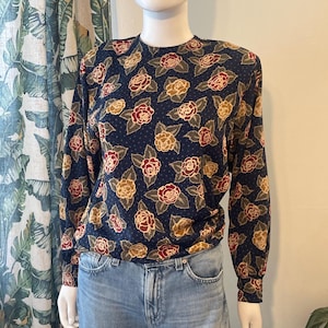 May include: A long-sleeved blouse with a navy blue background and a floral pattern of red, tan, and yellow roses. The blouse is paired with light blue denim jeans. The blouse has a round neckline and is made of a soft, flowing fabric.