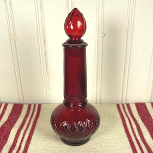 May include: A tall, ruby red glass decanter featuring a bulbous base with a diamond-cut pattern and a smooth, cylindrical neck. The decanter is topped with a matching red glass stopper shaped like a pointed, faceted bud or flame finial.