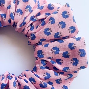 May include: A pink scrunchie with a blue floral pattern. The scrunchie is made of fabric and has a gathered look.