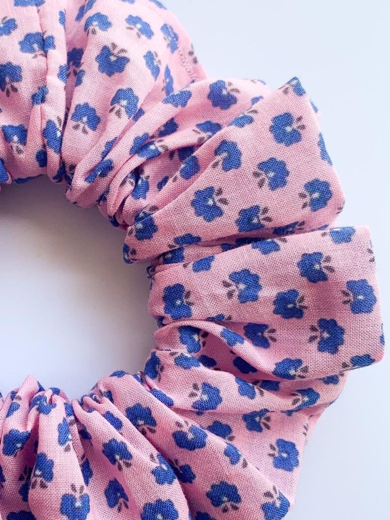 May include: A pink scrunchie with a blue floral pattern. The scrunchie is made of fabric and has a gathered look.