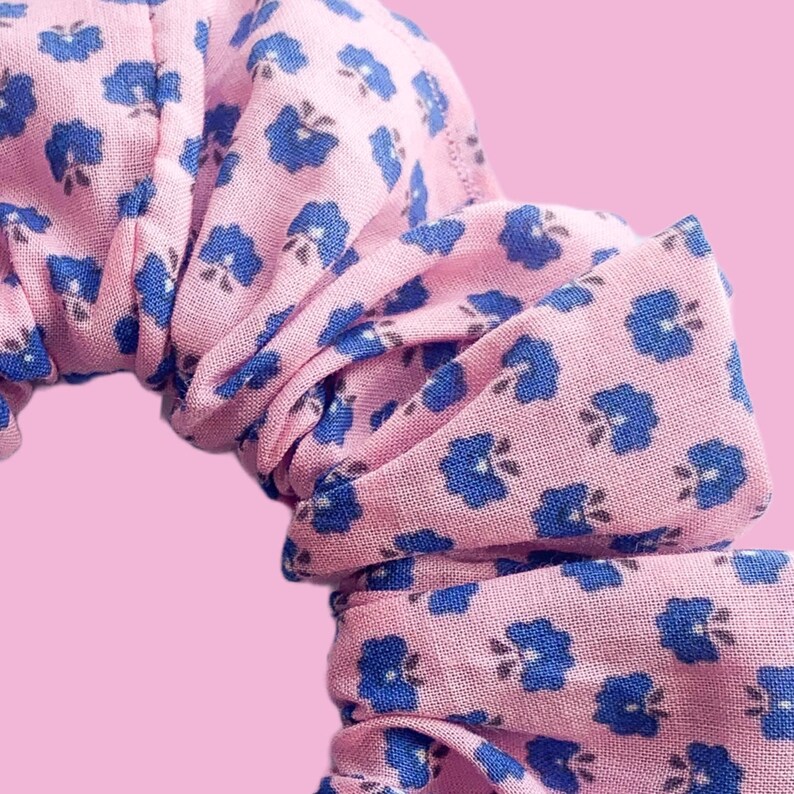 May include: A pink scrunchie with a blue floral pattern. The scrunchie is made of fabric and is gathered into a circle.