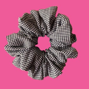 May include: A black and white gingham scrunchie, a hair accessory, set against a bright pink backdrop. The fabric is gathered and ruffled, forming a voluminous, circular shape. The classic pattern adds a touch of vintage charm.