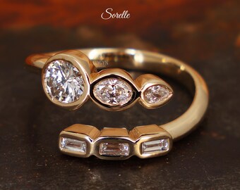May include: Gold-toned ring with multiple diamonds. The ring features a large, round diamond, two smaller oval diamonds, and three rectangular diamonds. The word "Sorelle" is written in cursive. The ring is from Elygyn Jewelry.