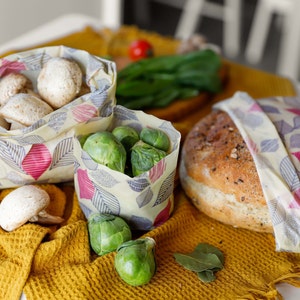 Peut inclure: Trois emballages alimentaires réutilisables avec un motif de feuilles dans des tons de rose, gris et jaune. Les emballages sont remplis de champignons, de choux de Bruxelles et d'un pain. Les emballages sont sur une surface en tissu jaune.
