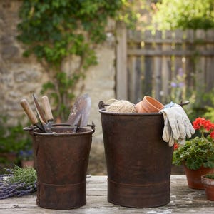 Può includere: Due secchi di metallo usurato su un tavolo di legno rustico, pieni di attrezzi da giardinaggio, spago, piccoli vasi di terracotta e guanti da giardinaggio. La scena è ambientata all'aperto con un muro di pietra e verde sullo sfondo.