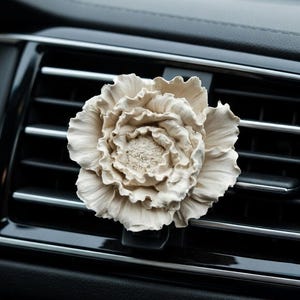 May include: A cream-coloured, flower-shaped car air freshener clipped to a black car vent. The air freshener has detailed petals and a textured centre, providing a decorative and aromatic element to the vehicle's interior.