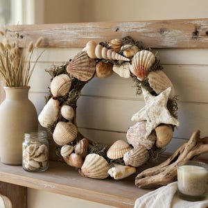 May include: A coastal-themed wreath made of various seashells, a starfish, and greenery. The wreath is displayed on a wooden shelf with a vase of dried flowers, a jar of shells, and a candle. The color palette is neutral with natural tones.