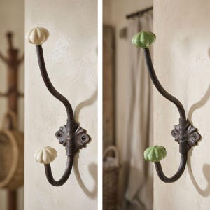 Könnte beinhalten: Zwei dekorative Wandhaken mit rustikalem Design. Jeder Haken hat einen dunklen, gebogenen Metallarm mit dekorativer Basis und einem Keramikknauf. Ein Haken hat einen cremefarbenen Knauf, der andere einen grünen Knauf.