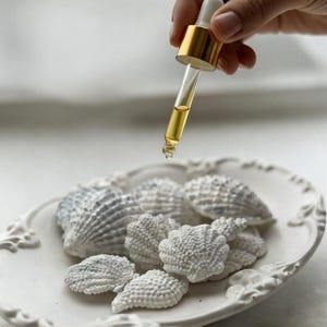 Op de afbeelding: Een goudkleurige druppelaar geeft een gouden vloeistof af over een wit, sierlijk, ovaalvormig bord gevuld met witte schelpvormige objecten. De schelpen hebben een textuur en zijn op het bord gerangschikt.