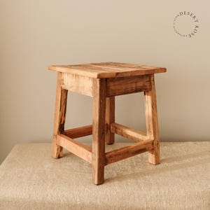 Könnte beinhalten: Ein rustikaler Holzhocker mit naturbelassenem Holzfinish. Der Hocker hat ein schlichtes Design mit vier Beinen und einer quadratischen Oberseite.