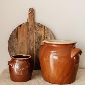 Puede incluir: Naturaleza muerta rústica con una tabla de cortar de madera y dos ollas de cerámica marrón con asas. Las ollas tienen un acabado brillante y un borde de color claro. La tabla de cortar es redonda y de madera reciclada.