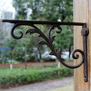 Può includere: Una staffa decorativa in ghisa marrone scuro con un ornato design a foglie a spirale. La staffa è montata su un palo di legno e presenta un braccio di supporto curvo e un ricciolo decorativo in basso. Progettata per appendere oggetti.
