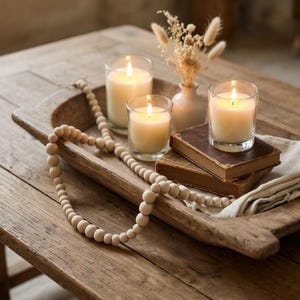 Könnte beinhalten: Ein Holztablett mit drei brennenden Kerzen in Glashaltern, einer kleinen Vase mit Trockenblumen und zwei antiken Büchern. Eine Perlenkette liegt über dem Tablett und dem Holztisch und erzeugt eine gemütliche, rustikale Ästhetik.