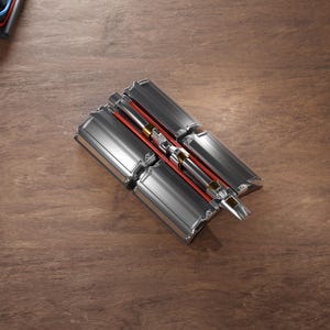 Puede incluir: Un dispositivo metálico plateado y rojo con múltiples paneles rectangulares y un componente cilíndrico central. El dispositivo está colocado sobre una superficie de madera, mostrando su intrincado diseño y elementos mecánicos.