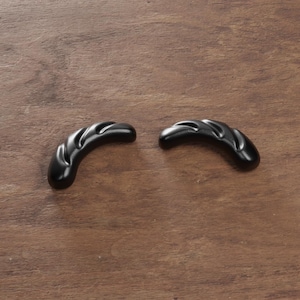 May include: Two black, curved cabinet pulls with a textured, ridged design. The hardware is made of a solid, dark material and is set against a warm, brown wooden surface. These pulls are likely for drawers or doors.