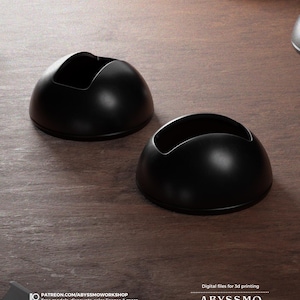 May include: Two black 3D printed objects with a rounded shape and a cutout in the centre. The objects are on a wooden surface.