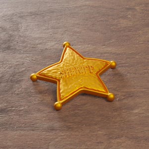 May include: A gold-colored sheriff's badge in the shape of a five-pointed star. The word "SHERIFF" is embossed across the center. The badge has rounded edges and small, spherical accents at each point. The badge is set against a wooden background.
