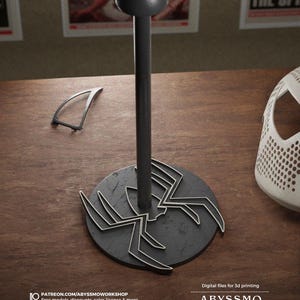 May include: A black Spider-Man helmet stand with a circular base featuring a spider design. The stand has a tall, slender pole and a rounded top. A white mask and a small, clear piece are also visible.