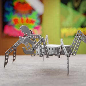 Op de afbeelding: Een zilveren metalen speelgoedspin met beweegbare poten en een lichaam van geperforeerde metalen platen. Het speelgoed is geconstrueerd met klinknagels en heeft een mechanische uitstraling. De achtergrond toont kleurrijke abstracte kunst.