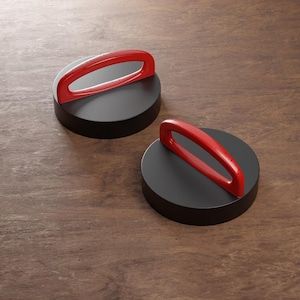 May include: Two black, circular objects with red, arched handles. The objects are on a wooden surface. The handles are a bright, glossy red, contrasting with the matte black of the circular bases. The design is simple and modern.