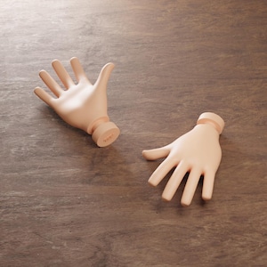 Op de afbeelding: Twee losstaande, lichtgekleurde handsculpturen met open handpalmen en een cilindrische basis. De handen zijn geplaatst op een bruin oppervlak met houtnerf. De sculpturen lijken gemaakt van een glad, mat materiaal.