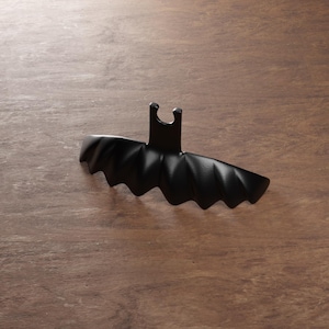Puede incluir: Un tapón de botella negro con forma de murciélago y diseño de alas festoneadas. El tapón tiene un pequeño asa rectangular y está hecho de un material mate. El tapón está diseñado para encajar en la parte superior de una botella.