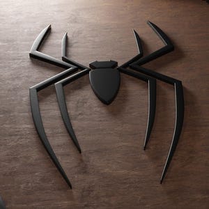 Puede incluir: Decoración de pared negra con forma de araña, con ocho patas largas y puntiagudas y un cuerpo en forma de escudo. La araña está hecha de un material negro mate y está sobre una superficie de madera marrón.