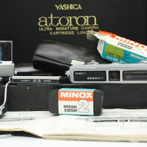 May include: A vintage Yashica Atoron ultra miniature camera kit. The kit includes a silver camera, film, flash, and accessories. The camera is labelled "Yashica Atoron" and "Minox". The kit is in a black box with the Yashica logo.