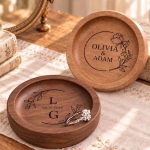 May include: Two round wooden ring dishes with engraved designs. The lighter dish features a floral circle with the text OLIVIA & ADAM. The darker walnut dish displays the initials L and G with the date 08.16.2024 and a silver ring inside.