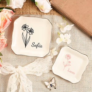 May include: Two off-white square trinket dishes with floral designs and names. One dish has a black floral design with the name "Sofia", and the other has a pink floral design with the name "Lucas". Both dishes have a gold trim.