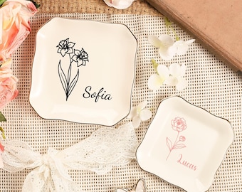 May include: Two off-white square trinket dishes with floral designs and names. One dish has a black floral design with the name "Sofia", and the other has a pink floral design with the name "Lucas". Both dishes have a gold trim.