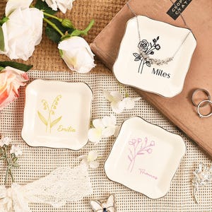 May include: Three off-white ceramic jewelry dishes with gold trim. Each dish features a floral design and a name: "Miles", "Emilia", and "Thomas". A silver necklace, rings, and a decorative butterfly are also present.
