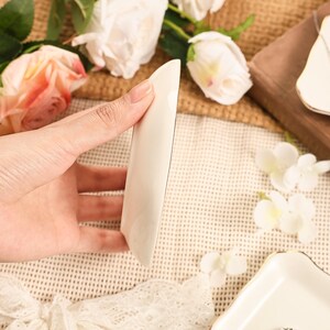 May include: A white ceramic serving dish with a curved, elongated design. The dish is held in a hand, showcasing its smooth, glossy surface. The background includes white flowers and a wooden board, suggesting a decorative or serving context.