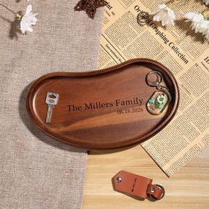 Op de afbeelding: Een houten boonvormig dienblad met de gegraveerde tekst "The Millers Family" en de datum "06.18.2026". De lade bevat een sleutel en een sleutelhanger met een bloemenontwerp. Een leren sleutelhanger is ook zichtbaar.
