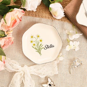May include: A cream-colored, irregularly shaped plate with a black rim. The plate features a floral design of yellow and white flowers with green stems and the name "Stella" in black script. The plate is surrounded by flowers and decorative items.