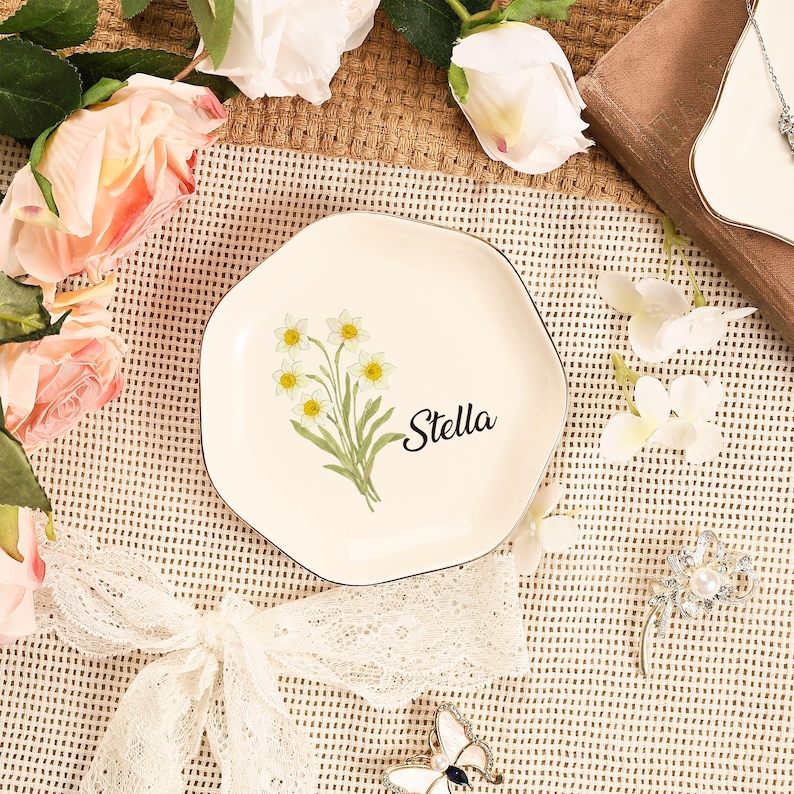 May include: A cream-colored, irregularly shaped plate with a black rim. The plate features a floral design of yellow and white flowers with green stems and the name "Stella" in black script. The plate is surrounded by flowers and decorative items.