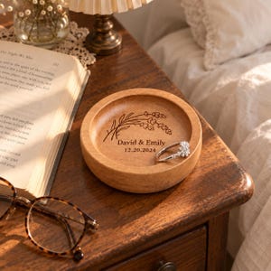 May include: A round wooden trinket dish featuring a laser-engraved lavender sprig, the names David & Emily, and the date 20.12.2024. A silver-toned ring with clear stones rests inside the bowl, which has a smooth, light brown wood grain finish.