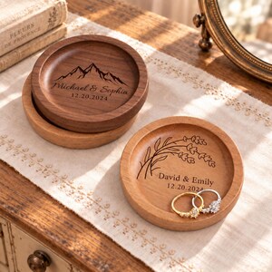May include: Two round wooden ring dishes. The darker dish features a mountain range engraving with the text Michael & Sophia 12.20.2024. The lighter dish features a lavender sprig engraving with the text David & Emily 12.20.2024 and two rings.