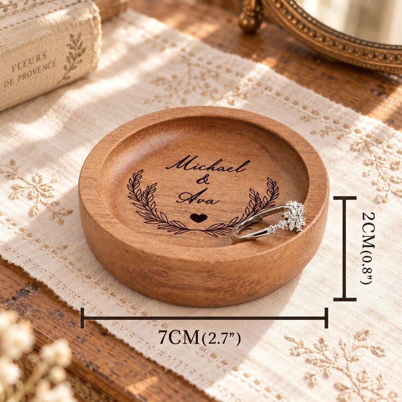 May include: A round wooden ring dish featuring the engraved names Michael & Ava with a laurel wreath and heart design. The dish measures 7 cm (2.7 inches) in diameter and 2 cm (0.8 inches) in height, with a silver-toned ring resting inside.