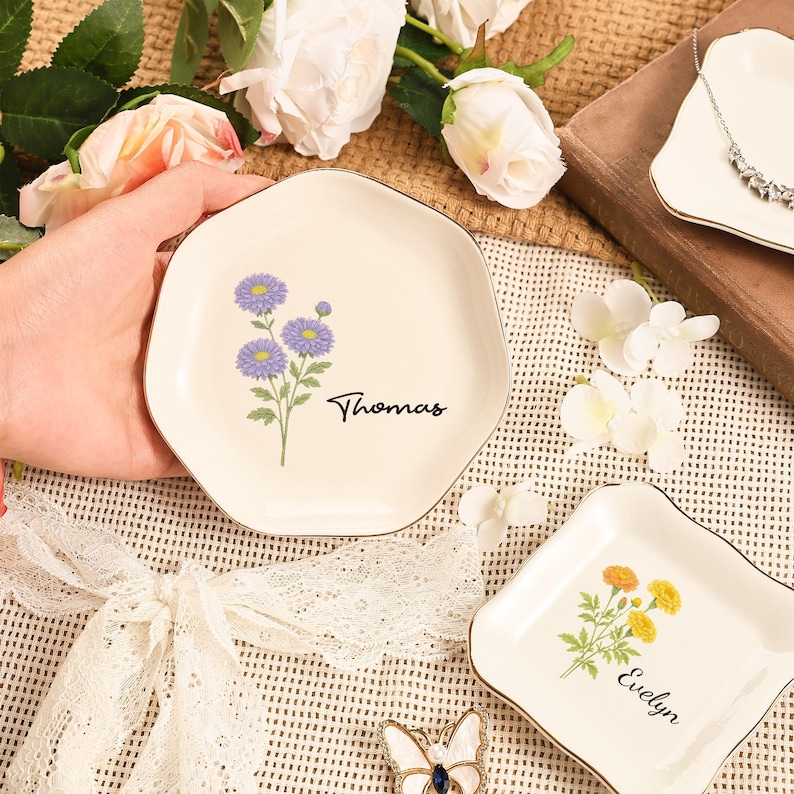 May include: Off-white ceramic trinket dish with a gold rim, featuring a purple floral design and the name "Thomas" in black script. Another dish with yellow flowers and the name "Evelyn" is also visible.
