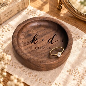 May include: A round, dark brown wooden ring dish featuring a smooth, polished finish. The center is engraved with the lowercase letters k + d in a cursive script, followed by the date 10.02.24. A gold-toned ring with a clear stone rests inside.