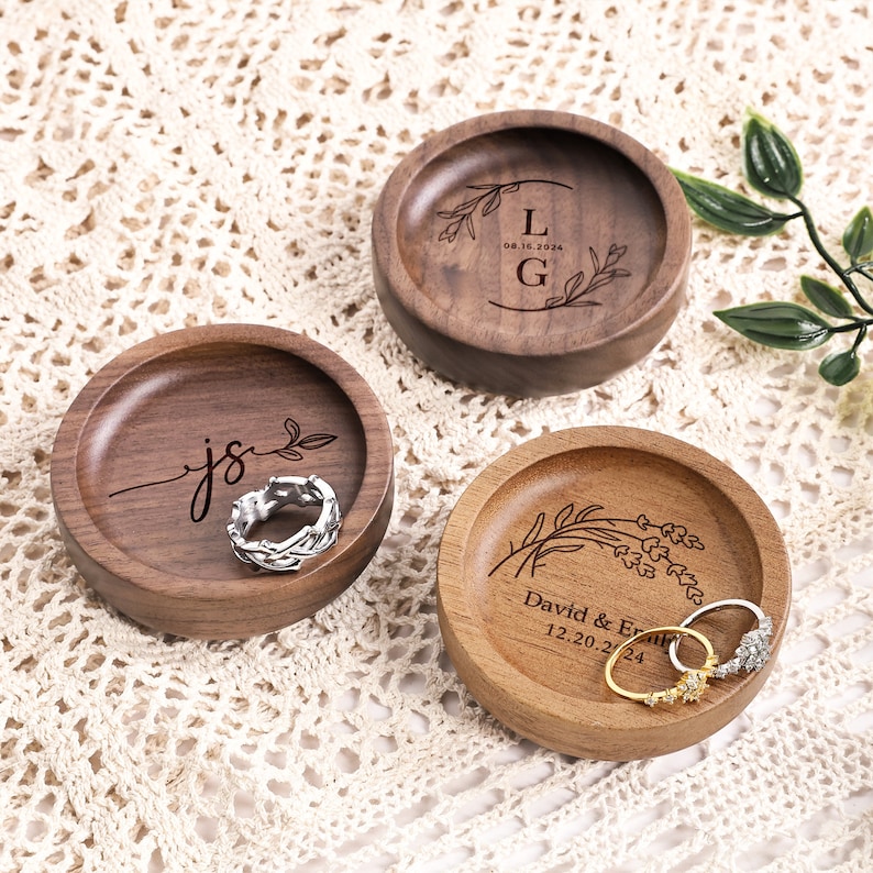 May include: Three round wooden ring dishes with engraved designs. One dish features the letters L and G with a date, another shows the letters js, and the third displays the names David & Emily with a date. Two dishes hold metal rings.